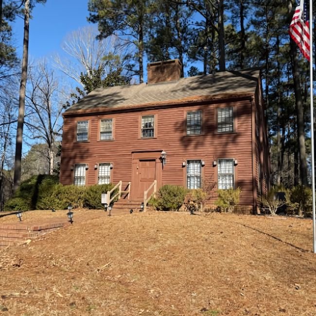 A two-story brown wooden house with a central chimney, six-pane windows, and a front door with steps. Surrounded by pine trees and an American flag, it's the kind of home Joe Buy Houses seeks for a fast cash offer.