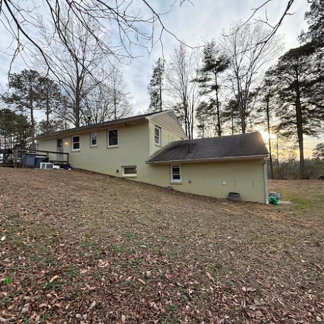 A yellow house with a sloped yard sits among tall leafless trees on a cloudy day. Fallen leaves cover the ground, and the sun sets behind the trees—an inviting scene for Joe Buy Houses to find your next home.