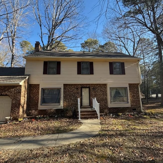 A two-story home with beige siding and brick on the lower level, featuring a central front door with white steps and railings, two large front windows, and a leaf-covered yard under bare trees—perfect for a quick cash offer from Joe Buy Houses.