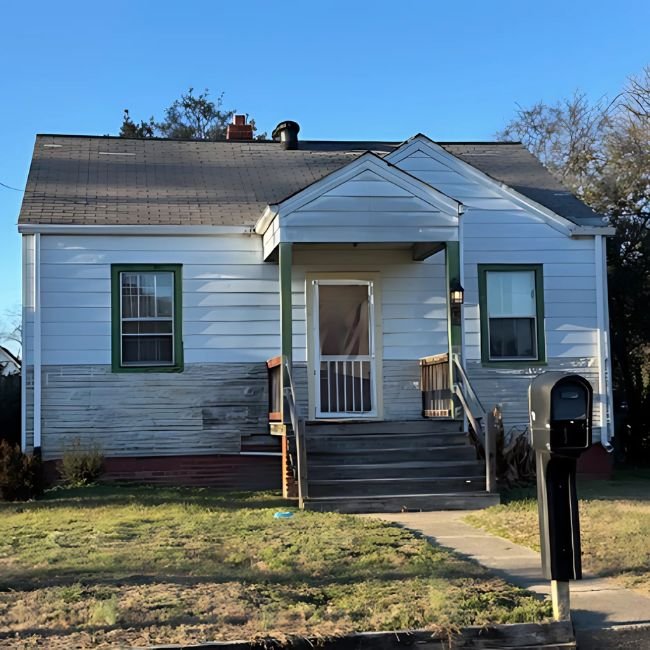 A small, single-story house with white siding, green trim, a front porch with steps, and a black mailbox by the sidewalk. Joe Buy Houses can make a cash offer for houses like this with its patchy yard and clear blue sky.