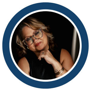 A woman with shoulder-length wavy hair and glasses smiles softly, resting her chin on her hand. She wears a sleeveless top, bracelets, and sits against a dark background framed by a blue and white circular "Joe Buy Houses" border.